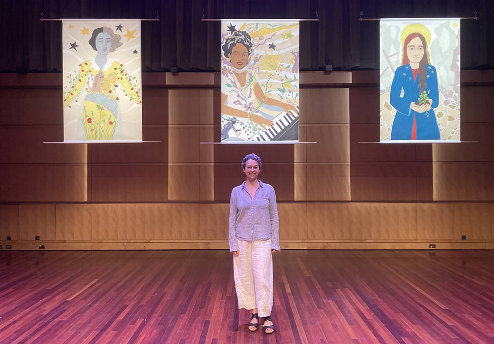 Milly Charles standing under 3 pieces of her work.